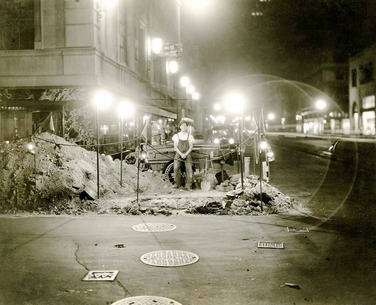 Black and white photo of a Con Edison worker standing in front of a work site dug into a New York City street corner.