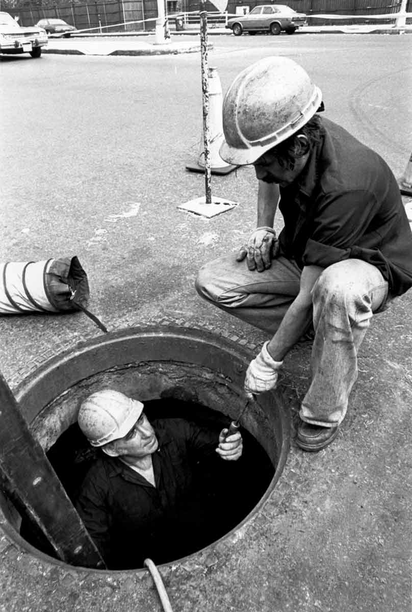 Black and white photo of a Con Edison worker handing a tool to a colleague who is inside a manhole.