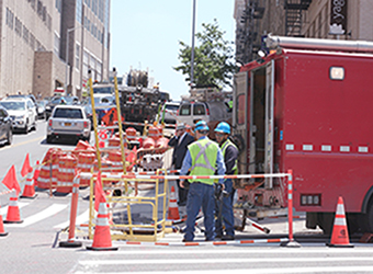 Con Edison workers surrounded by safety cones, are working on a city street.