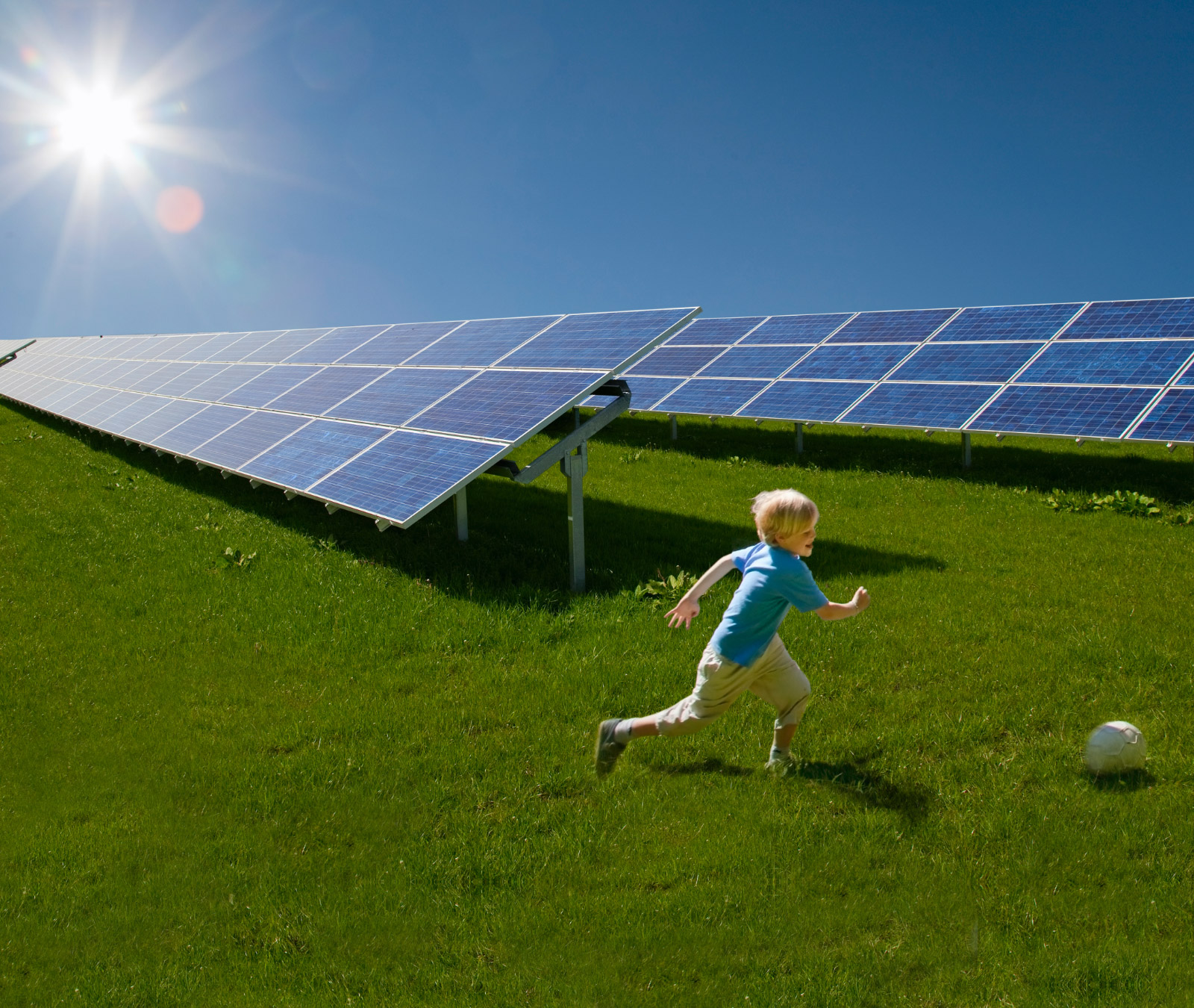 A child is chasing a soccer ball in a grassy field with solar panels in the background.